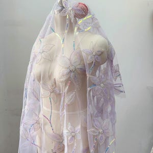 May include: A sheer, light purple tulle fabric draped over a mannequin, featuring embroidered floral motifs with iridescent sequins and small white pearl-like beads at the center of each flower, creating a delicate, layered, and shimmering appearance.