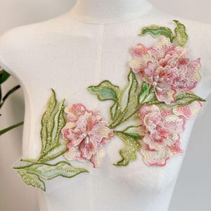Könnte beinhalten: Eine florale Spitzenapplikation mit gestickten rosa und weißen Blumen sowie grünen Blättern. Das Design ist mit kleinen weißen und rosa Perlen sowie Pailletten verziert, was eine strukturierte Oberfläche auf einem leichten Netzstoff erzeugt.