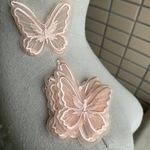 May include: Two light pink butterfly appliques. The butterflies are made of sheer fabric with embroidered details, and are layered for a 3D effect. The wings have a subtle shimmer, and the edges are outlined with a matching thread.