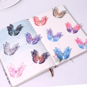 May include: A journal open to two pages, adorned with several colorful butterfly stickers. The butterflies feature a variety of colors, including pink, purple, blue, and brown, with a small gem in the center of each. The journal has a brown leather bookmark.