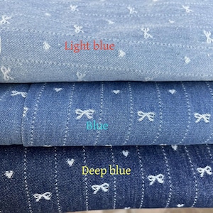 Puede incluir: Tres muestras de tela vaquera con corazones y lazos bordados en blanco. La muestra superior es azul claro, la muestra del medio es azul y la muestra inferior es azul oscuro.