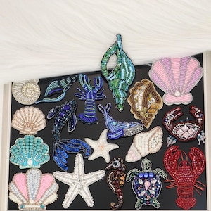 May include: A collection of embroidered patches featuring various marine life and seashells. The set includes a blue lobster, a red lobster, a sea turtle, a whale, a fish, two starfish, and several shells in pink, white, turquoise, and gold.