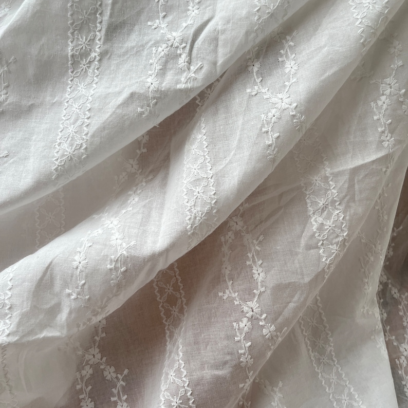 May include: White cotton fabric with delicate embroidered floral patterns. The fabric is folded and draped, showcasing the intricate details of the embroidery.