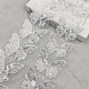 Pode incluir: Uma fita de renda branca com lantejoulas prateadas em forma de borboletas. A fita é perfeita para adicionar um toque de elegância a qualquer projeto.