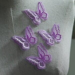 May include: Four lavender butterfly appliques with white embroidered details. The butterflies are made of a sheer, layered fabric, creating a three-dimensional effect. These decorative appliques are ideal for embellishing clothing, accessories, or craft projects.