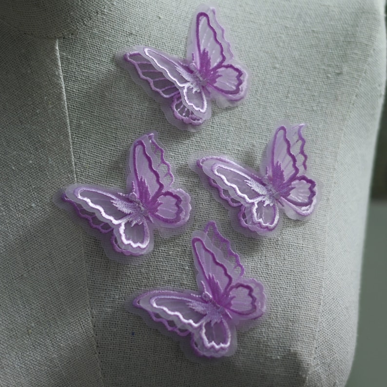 May include: Four lavender butterfly appliques with white embroidered details. The butterflies are made of a sheer, layered fabric, creating a three-dimensional effect. These decorative appliques are ideal for embellishing clothing, accessories, or craft projects.
