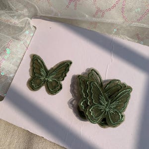 May include: Two green embroidered butterfly appliques. One is a single butterfly, and the other is a stack of three butterflies. The appliques are made of a sheer fabric with detailed stitching, ideal for crafting or embellishing projects.