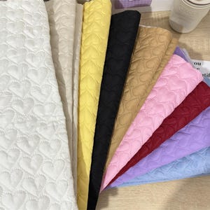 Op de afbeelding: Een verzameling gewatteerde stofstalen in verschillende kleuren, waaronder wit, beige, geel, zwart, bruin, roze, rood, paars en blauw. Elke stof heeft een hartvormig stikselpatroon, waardoor een textuur ontstaat. De stoffen zijn in een waaierachtige weergave gerangschikt.