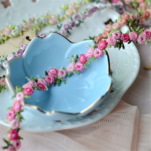 May include: A close-up of a delicate floral trim with pink and white flowers and green leaves. The trim is displayed on a light blue ceramic dish.