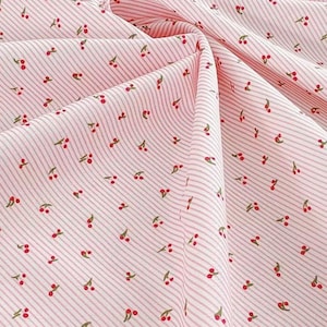 May include: Pink fabric featuring a pattern of small red cherries with green stems. The fabric has a striped design with thin, light pink lines. The fabric is folded, showing the texture and pattern of the material.