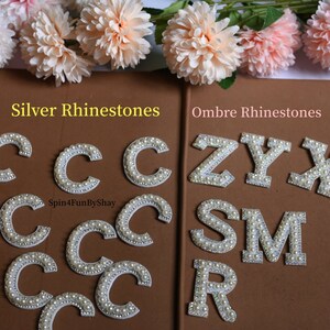May include: Two sets of iron-on letters decorated with white pearl-like beads. The letters on the left are all "C" and are labeled "Silver Rhinestones". The letters on the right are "Z", "Y", "X", "S", "M", and "R" and are labeled "Ombre Rhinestones".
