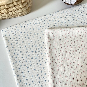 Op de afbeelding: Twee stukken witte geribbelde stof met kleine bloemenprints. Het ene stuk heeft een patroon van kleine blauwe bloemetjes, terwijl het andere stuk kleine roze bloemetjes met groene blaadjes bevat, beide gerangschikt in verticale rijen.