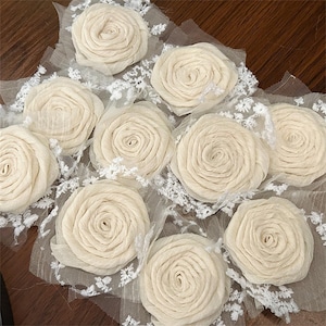 May include: A collection of off-white fabric roses arranged on a sheer, light-coloured mesh base. Each rose features layered, spiraled petals. The mesh background is accented with delicate, white floral lace trim detailing between the flowers.