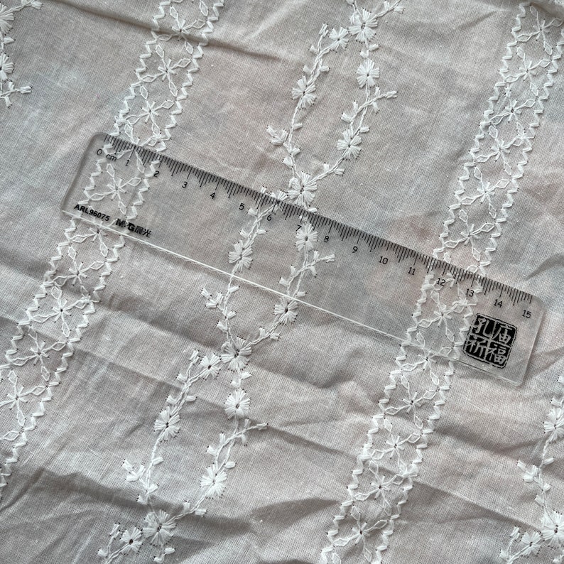 May include: White cotton fabric with a delicate embroidered pattern of small flowers and vines. A clear ruler with black markings is placed on the fabric, measuring 15 centimeters.