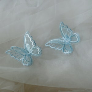May include: Two pale blue embroidered butterfly appliques with white detailing. The butterflies are set against a soft, white, sheer fabric. The wings feature intricate stitching, creating a delicate and airy design.