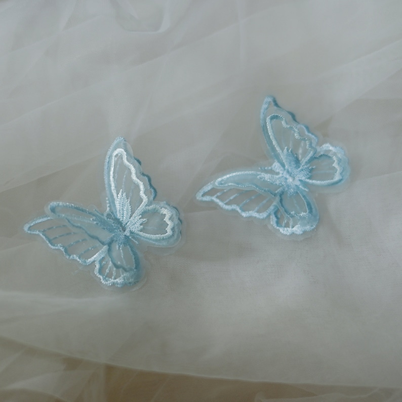 May include: Two pale blue embroidered butterfly appliques with white detailing. The butterflies are set against a soft, white, sheer fabric. The wings feature intricate stitching, creating a delicate and airy design.
