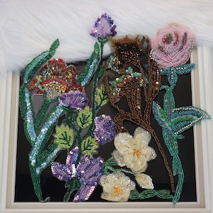 May include: A decorative floral appliqué featuring intricate beadwork and sequins in shades of pink, purple, green, brown, and cream. The design showcases various flower stems and leaves crafted with detailed embroidery on a sheer mesh base.