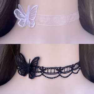 May include: Two chokers are shown. The top one features a white butterfly and a pale pink ribbon. The bottom choker is black lace with a black butterfly. Both are elegant and delicate accessories.