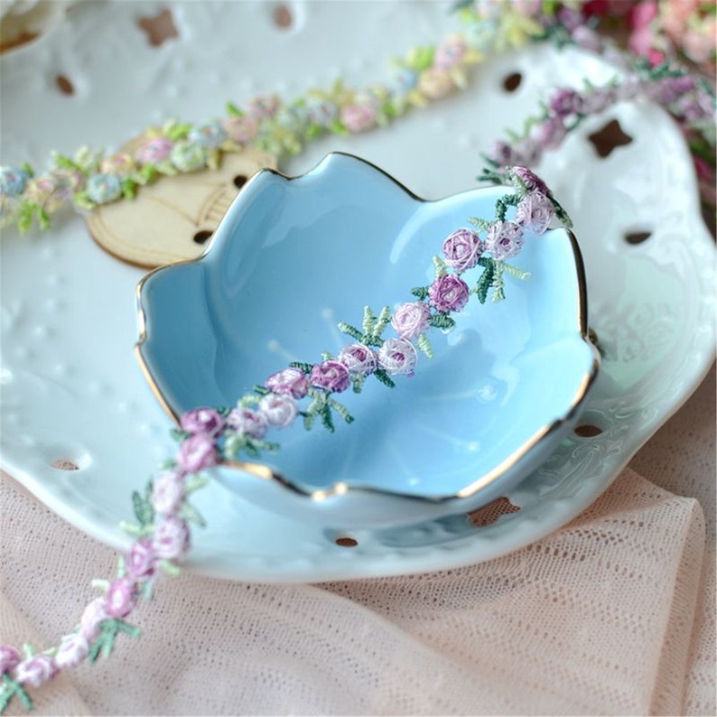 May include: A delicate floral trim with small purple and white flowers and green leaves. The trim is displayed on a light blue ceramic dish.