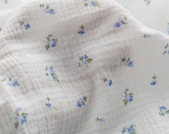 Op de afbeelding: Witte katoenen stof met een klein blauw bloemenpatroon. De stof heeft een textuur en is licht gekreukt.