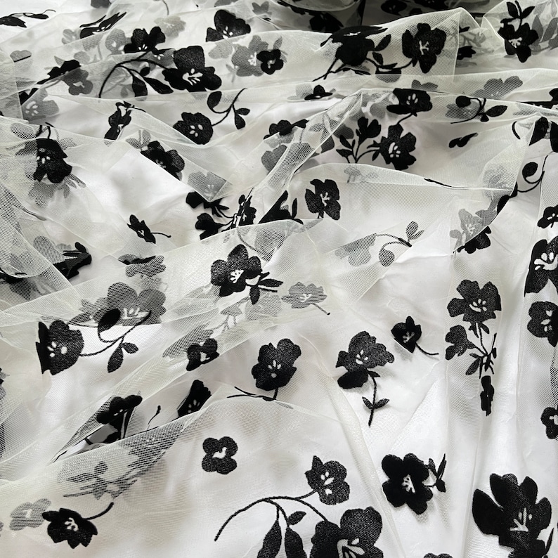 May include: White tulle fabric with black floral embroidery. The fabric is sheer and delicate, with a soft, flowing texture.