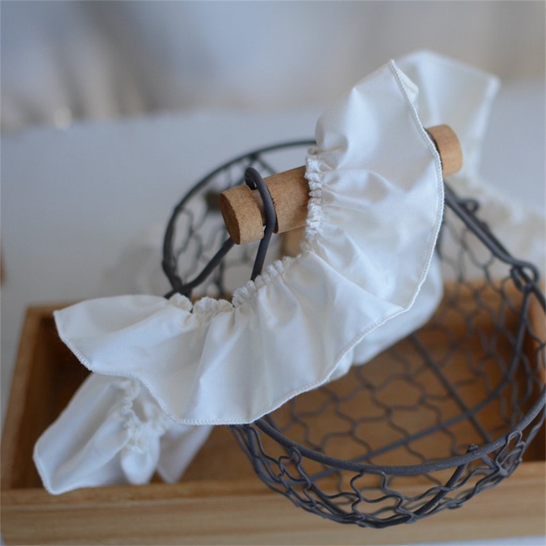 May include: A close-up of a white fabric ruffle detail, possibly a decorative element for a basket or other item. The ruffle is gathered and attached to a wooden dowel, with a wire basket in the background.