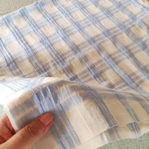 Peut inclure: Gros plan sur un tissu à carreaux bleu clair et blanc. Le tissu a un aspect doux et texturé avec un motif quadrillé. Le tissu est plié, révélant les couches et la texture du matériau.