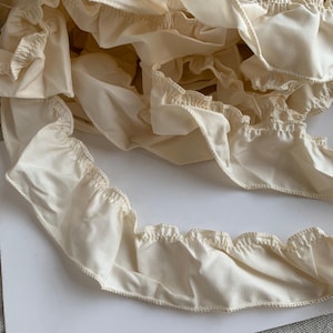 May include: Close-up of ivory-colored fabric ruffles. The ruffles have a scalloped edge and are gathered along a seam. The fabric appears to be a soft, lightweight material, possibly cotton or linen. The ruffles are layered and create a textured effect.