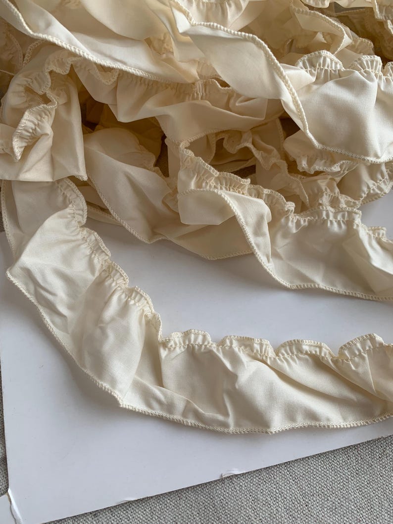May include: Close-up of ivory-colored fabric ruffles. The ruffles have a scalloped edge and are gathered along a seam. The fabric appears to be a soft, lightweight material, possibly cotton or linen. The ruffles are layered and create a textured effect.