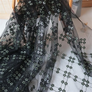 May include: Black sheer fabric with a floral pattern. The fabric is decorated with small, silver sequins that reflect light. The floral design is evenly spaced, creating a delicate and elegant look. Suitable for dresses and other apparel.