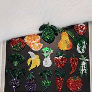 Op de afbeelding: Een verzameling patches in de vorm van groenten en fruit, versierd met kleurrijke kralen en pailletten. Het assortiment bevat ontwerpen van een limoen, peer, aardbei, broccoli, sinaasappelschijfjes, banaan, chilipeper, aubergine, radijs en tomaat.