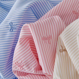 May include: Three fabric swatches with white stripes on a blue, pink, and beige background. Each swatch has a small embroidered bow in a matching color.