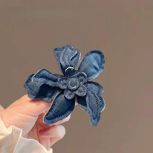 Può includere: Un fermaglio per capelli a forma di fiore in denim con un design a petali a strati. Il fiore è di colore blu scuro con dettagli di cucitura bianchi. L'accessorio per capelli è tenuto in mano su uno sfondo neutro.