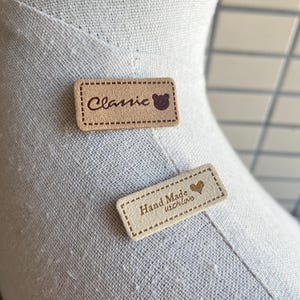 May include: Two rectangular, tan-colored fabric labels. One reads "Classic" with a bear icon, the other reads "Hand Made with love" with a heart icon. Both have a stitched border.