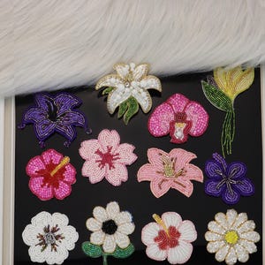 May include: A collection of twelve assorted flower-shaped patches crafted with intricate beadwork. The set includes various floral designs in vibrant colors such as white, purple, pink, yellow, and red, featuring detailed petal textures and centers.