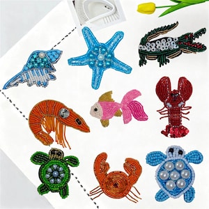 Pode incluir: Uma coleção de nove emblemas bordados com várias criaturas marinhas, incluindo uma estrela-do-mar azul, uma tartaruga verde, uma lagosta vermelha, um camarão laranja, um peixe rosa, um crocodilo verde, uma concha azul e um caranguejo laranja.