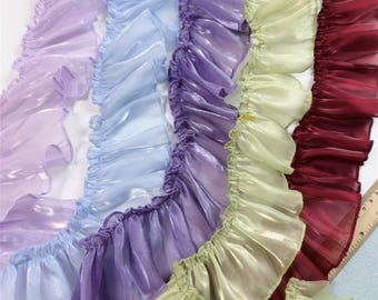 Puede incluir: Primer plano de varias filas de tela con volantes en tonos lavanda, azul claro, morado, verde pálido y burdeos. La tela tiene una calidad brillante e iridiscente, y los volantes están densamente reunidos.