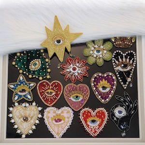 Puede incluir: Una colección de parches bordados y con cuentas que presentan varias formas, incluyendo corazones, estrellas, flores y una mano. Cada parche incorpora un motivo de ojo central representado en diversos colores, texturas y detalles.