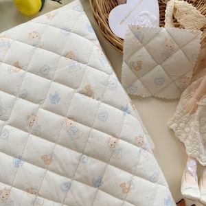 May include: A quilted fabric featuring a repeating pattern of small teddy bears in light brown, blue bows, and hearts on an off-white background. The diamond-stitched material has a soft texture and includes a matching square fabric swatch.