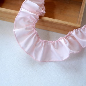 May include: A length of pale pink ruffled fabric, possibly satin or silk, with a gathered edge. The fabric has a soft, flowing appearance and a delicate, feminine aesthetic. The ruffled trim is ideal for sewing and craft projects.