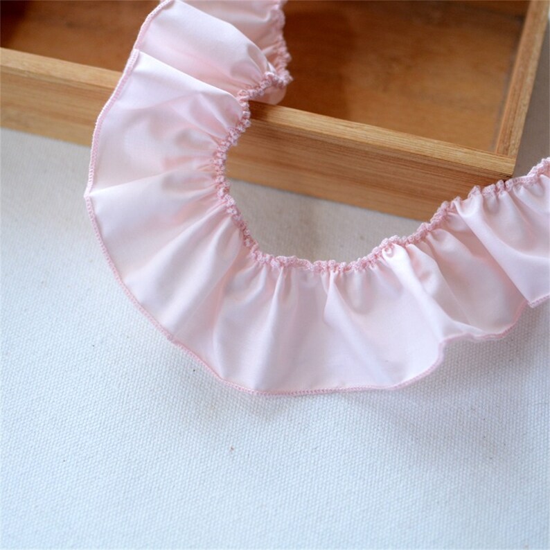 May include: A length of pale pink ruffled fabric, possibly satin or silk, with a gathered edge. The fabric has a soft, flowing appearance and a delicate, feminine aesthetic. The ruffled trim is ideal for sewing and craft projects.