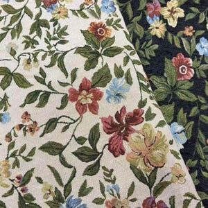 Op de afbeelding: Een geweven stof met een bloemenpatroon met rode, gele en blauwe bloemen tegen groene bladeren. Het textiel wordt in twee delen getoond, één met een crèmekleurige achtergrond en de andere met een donkerzwarte achtergrond.