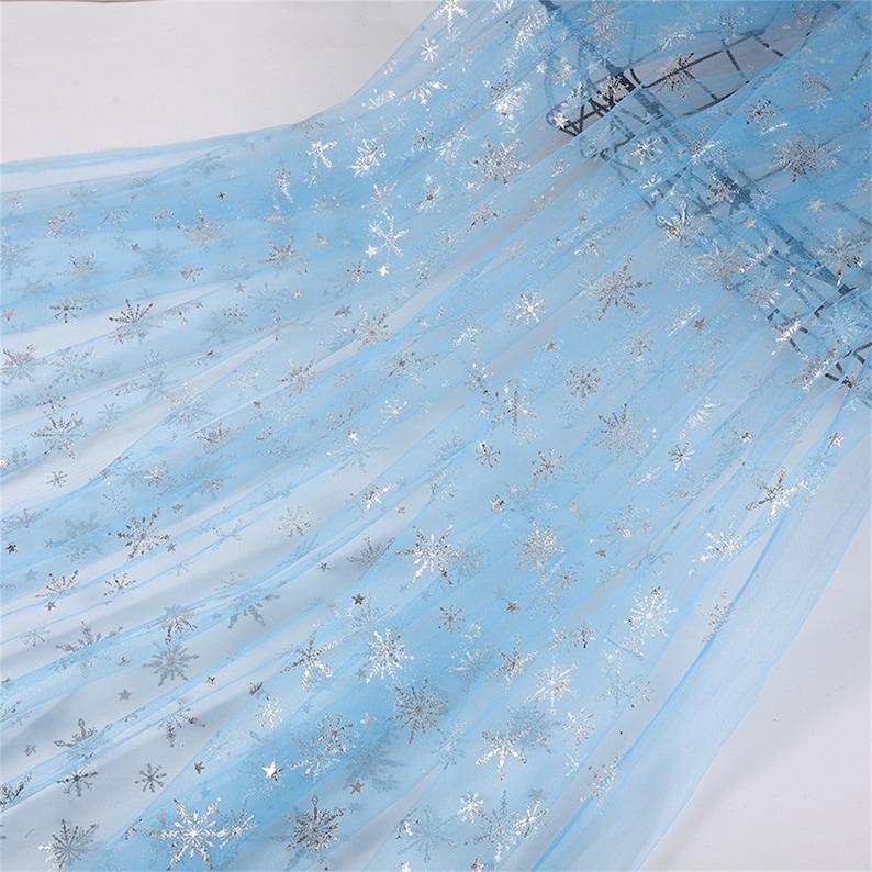 May include: Light blue tulle fabric with silver snowflake pattern. The fabric is sheer and delicate, perfect for crafting or decorating.