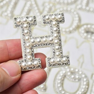 May include: A white felt letter H with a pearl and rhinestone embellishment. The letter is held in a hand.