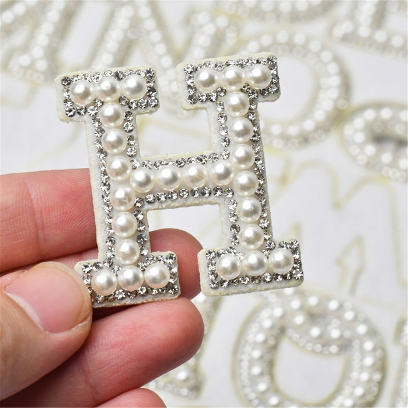 May include: A white felt letter H with a pearl and rhinestone embellishment. The letter is held in a hand.