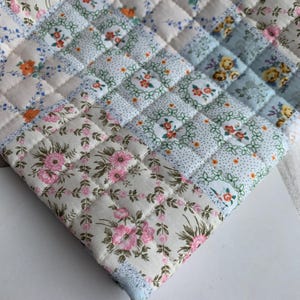 May include: A quilted fabric piece featuring a patchwork design of various floral and dotted patterns in shades of pink, blue, green, orange, and white. The textile is constructed with a grid of square stitches, creating a textured surface.