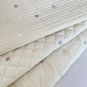 Può includere: Un tessuto trapuntato color crema con un motivo di piccoli fiori ricamati nei toni del rosa, blu e giallo. Il tessuto presenta due stili di cucitura distinti: uno con linee parallele orizzontali e uno con un motivo a losanghe.