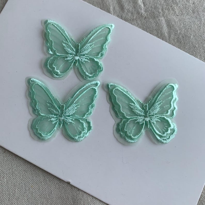 May include: Three mint green embroidered butterfly appliques. The butterflies feature detailed wing patterns and are arranged on a white surface. These appliques are suitable for crafts and embellishments.
