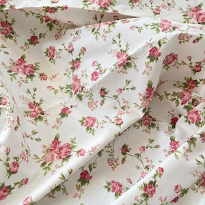 May include: A white fabric featuring a repeating floral pattern of small pink roses with green leaves and stems. The material is draped in soft folds, showcasing the continuous botanical print across the surface of the textile.