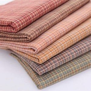 May include: A stack of folded fabric pieces featuring a consistent small-scale plaid pattern. The layers display a gradient of colours including deep red, warm brown, soft orange, muted grey, and a subtle greenish-blue hue with thin lines.